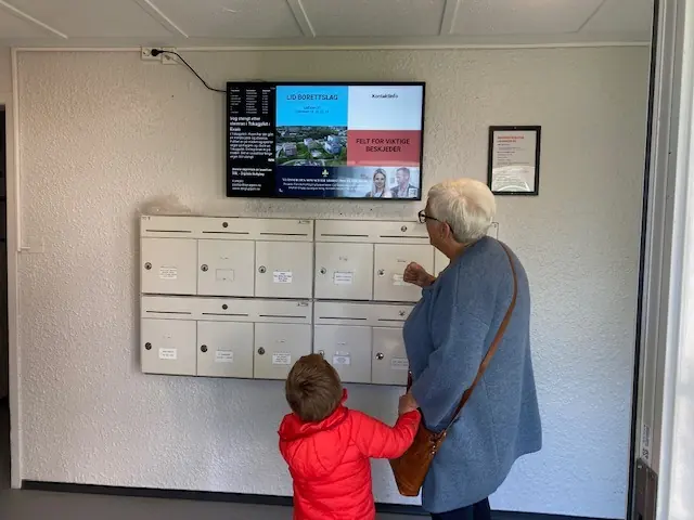 Bestemor og barn ved postkassene, med infoskjerm over